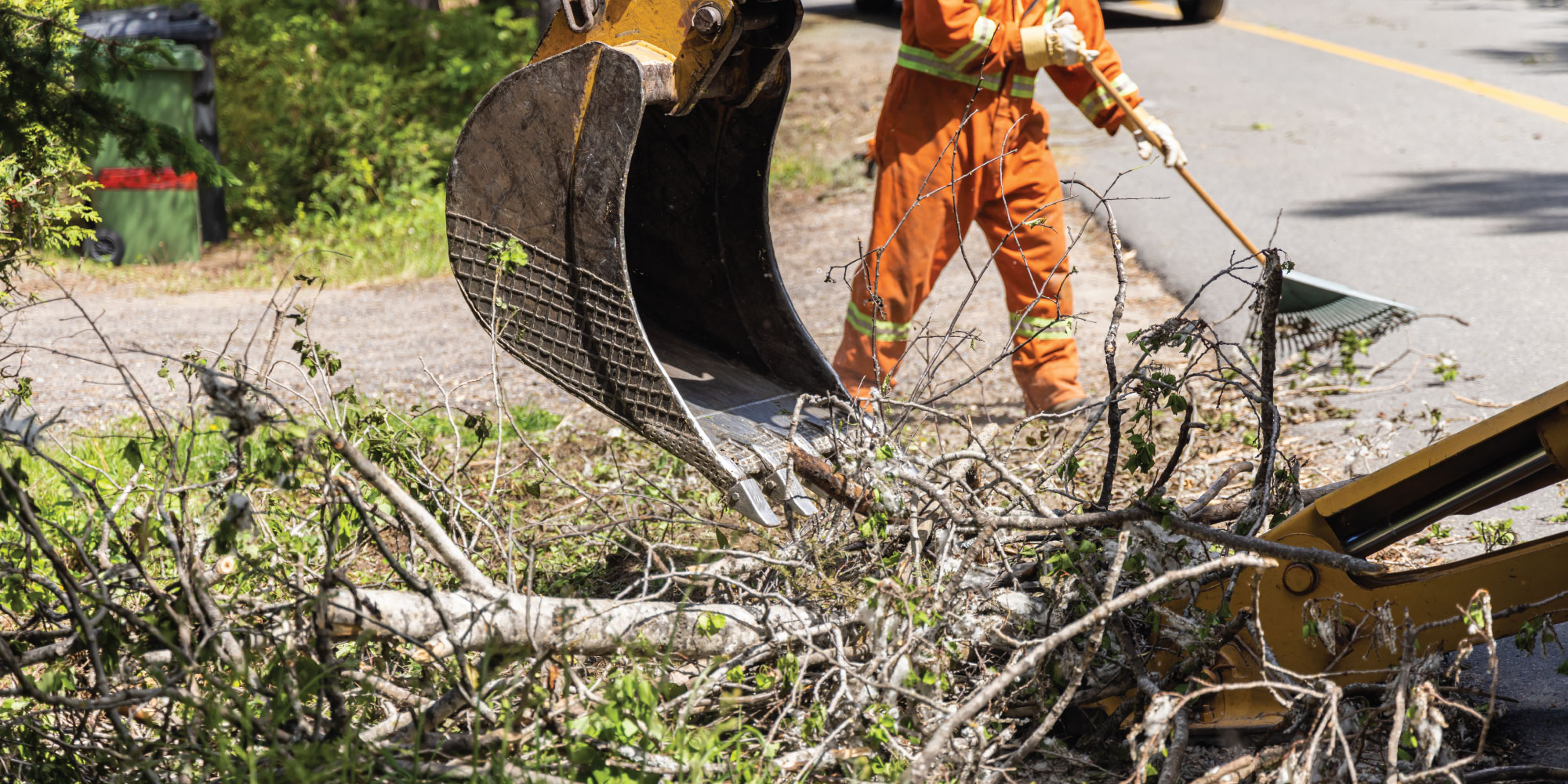 The bucket of an excavator picking up tree debris on the side of the road with a worker in an orange high-visibility jumpsuit raking in the background